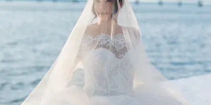 A bride in a white lace wedding dress and veil sitting by the water.