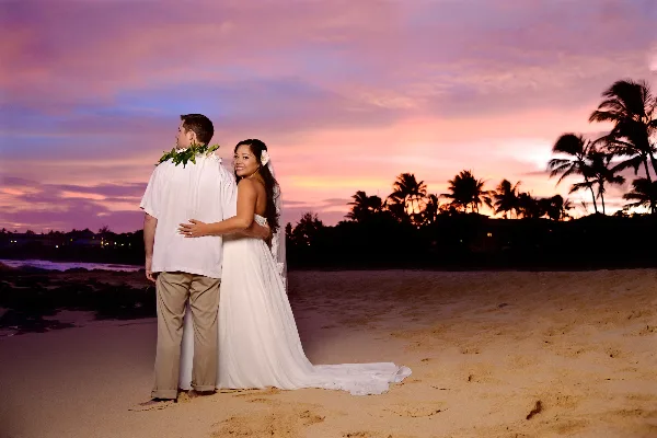 A bride and groom standing on a sandy beach at sunset with palm trees in the background.