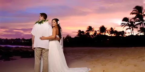 A bride and groom standing on a sandy beach at sunset with palm trees in the background.