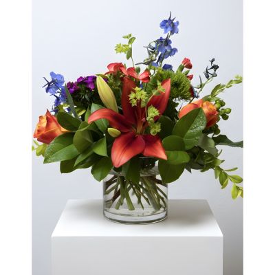 A vibrant floral arrangement featuring red lilies, purple flowers, and greenery in a clear glass vase on a white pedestal.