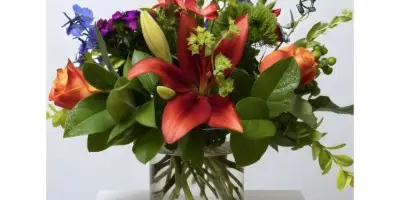 A vibrant floral arrangement featuring red lilies, purple flowers, and greenery in a clear glass vase on a white pedestal.