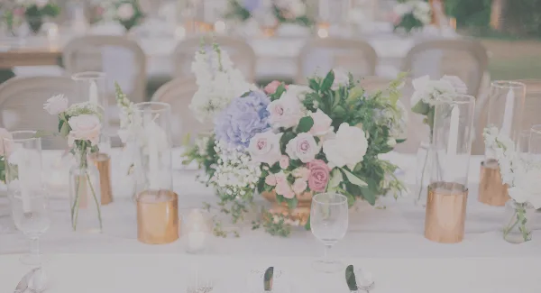 A wedding reception table decorated with a large floral centerpiece featuring pink and white roses, hydrangeas, and greenery, surrounded by glass candle holders and place settings.