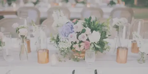 A wedding reception table decorated with a large floral centerpiece featuring pink and white roses, hydrangeas, and greenery, surrounded by glass candle holders and place settings.