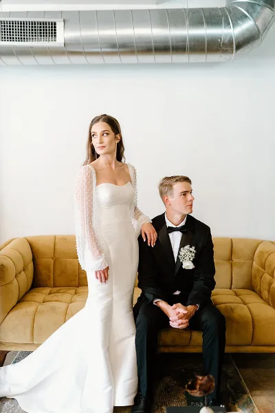 A bride in a white gown and a groom in a black tuxedo sitting on a mustard yellow sofa.