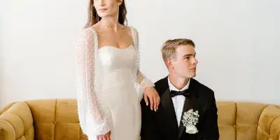 A bride in a white gown and a groom in a black tuxedo sitting on a mustard yellow sofa.