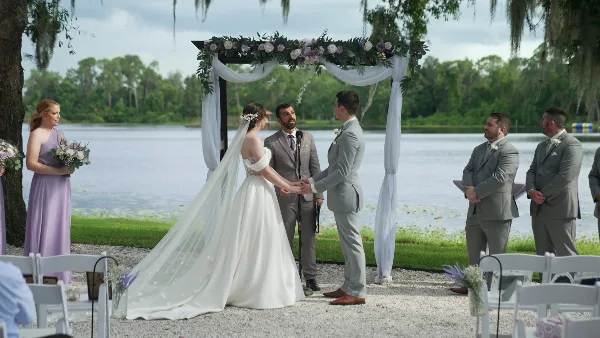 A bride and groom exchanging vows at a wedding ceremony by a lake, with an officiant and wedding party present.