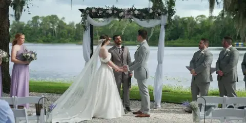 A bride and groom exchanging vows at a wedding ceremony by a lake, with an officiant and wedding party present.