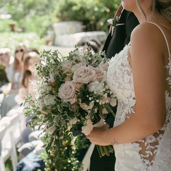 A bride in a white lace dress holding a large bouquet of blush roses and greenery during an outdoor wedding ceremony.
