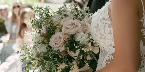 A bride in a white lace dress holding a large bouquet of blush roses and greenery during an outdoor wedding ceremony.