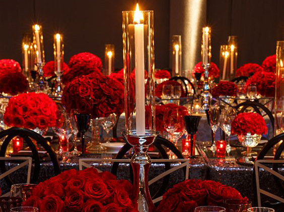 A wedding reception table decorated with lush red rose centerpieces and tall glass candle holders.