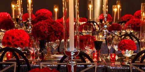 A wedding reception table decorated with lush red rose centerpieces and tall glass candle holders.