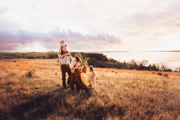 A family of six posing in a golden grassy field near a body of water during sunset.