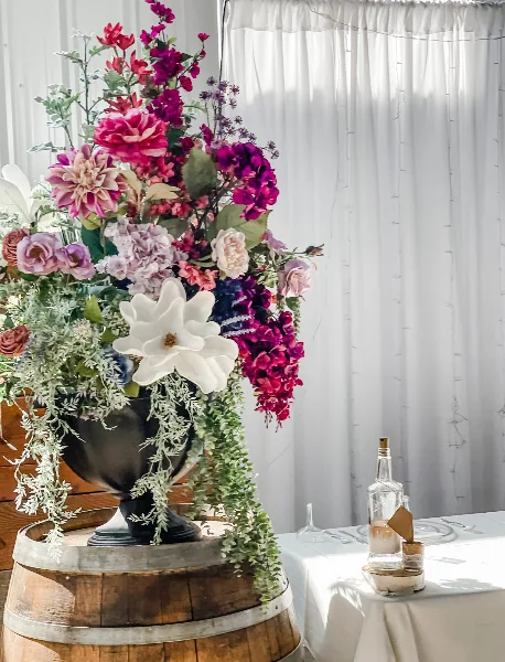 A tall, colorful floral centerpiece with pink and purple flowers in a silver urn, placed on a wooden barrel next to a white table setting.