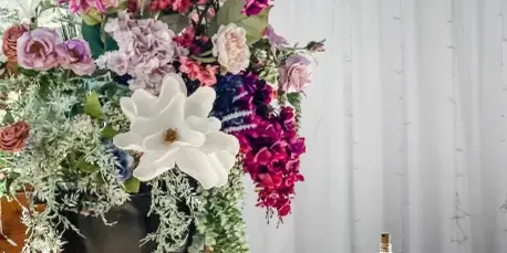 A tall, colorful floral centerpiece with pink and purple flowers in a silver urn, placed on a wooden barrel next to a white table setting.