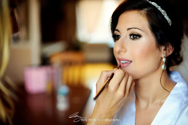 A makeup artist applying lip liner to a bride wearing a tiara and white robe.