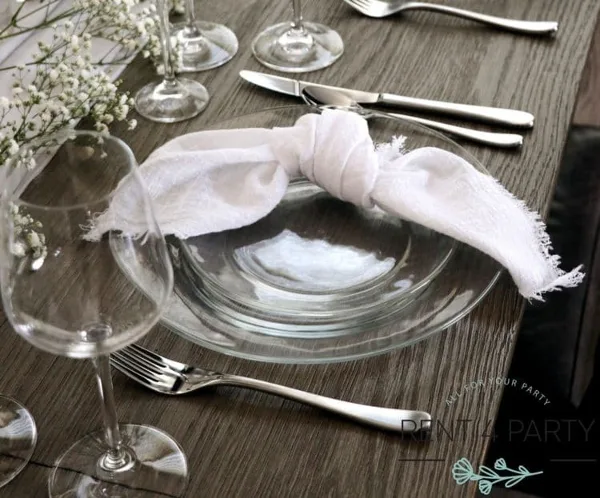 A close-up of a table setting with a clear glass plate, silver flatware, a white napkin, and a wine glass on a wooden table.