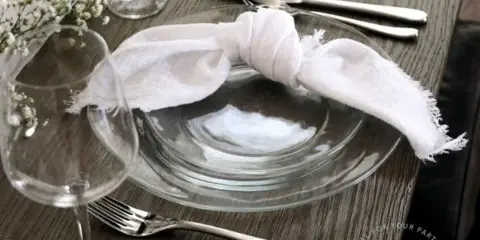 A close-up of a table setting with a clear glass plate, silver flatware, a white napkin, and a wine glass on a wooden table.