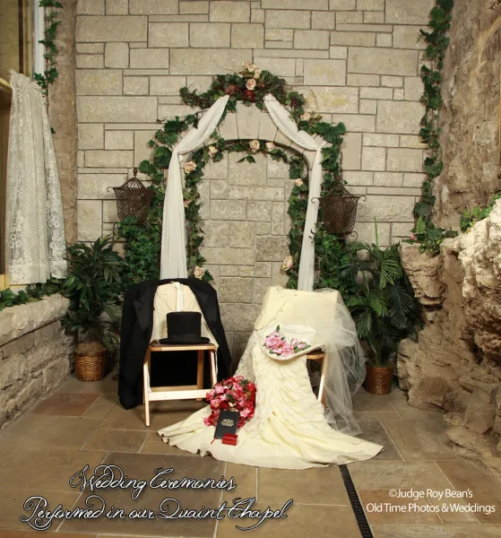 A wedding arch decorated with flowers and white fabric in a stone chapel, with a wedding dress and groom's suit displayed on chairs.