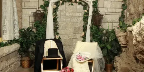 A wedding arch decorated with flowers and white fabric in a stone chapel, with a wedding dress and groom's suit displayed on chairs.
