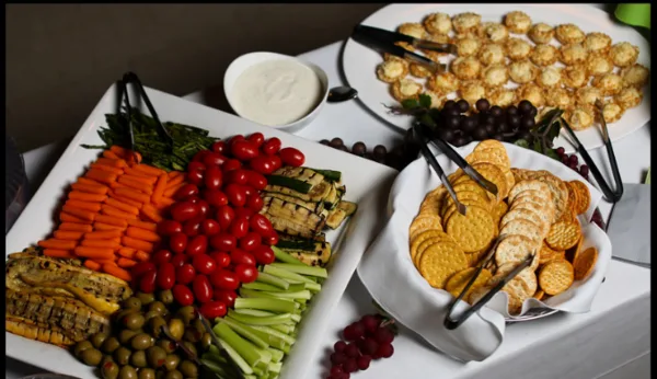 A catering table featuring a vegetable tray with carrots, celery, tomatoes, and olives, a bowl of dip, a basket of crackers, and a platter of small appetizers.