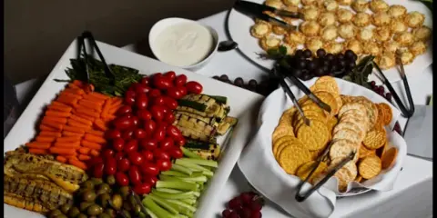 A catering table featuring a vegetable tray with carrots, celery, tomatoes, and olives, a bowl of dip, a basket of crackers, and a platter of small appetizers.