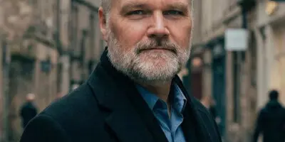 Portrait of Ryan Hinman, a man with a grey beard wearing a dark coat, standing on a city street.
