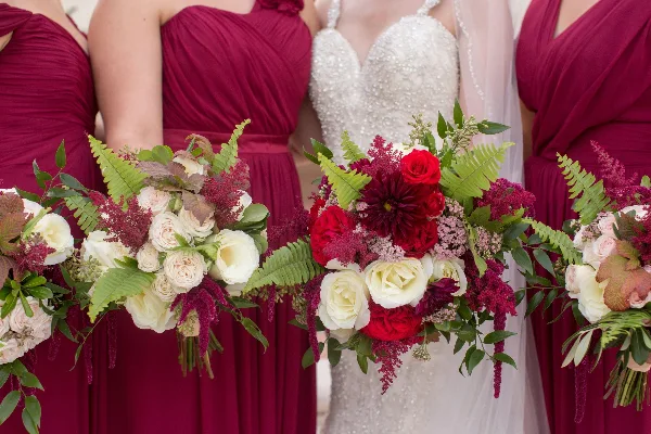 A bride in a white beaded dress and bridesmaids in maroon dresses holding floral bouquets.
