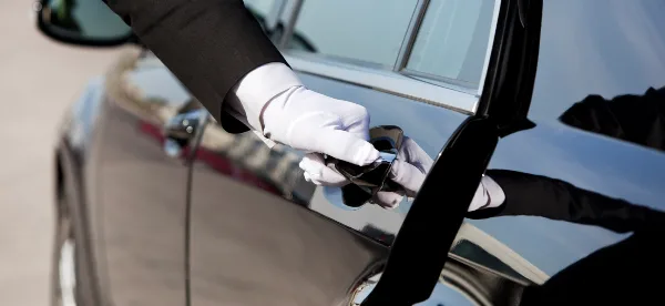 A chauffeur wearing a black tuxedo and white gloves opens the door of a black luxury car.