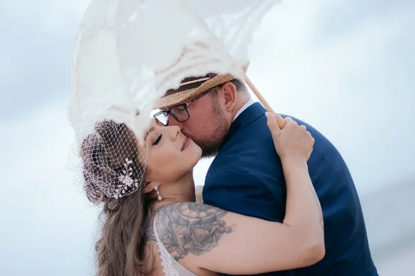 A bride with styled hair and makeup kissing a groom under a white parasol.