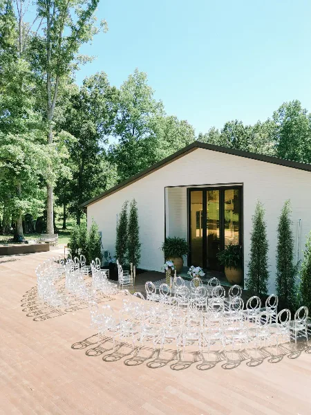 Outdoor wedding ceremony setup with rows of clear chairs on a wooden deck in front of a white Pavilion building.