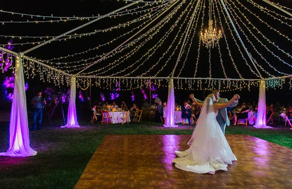 A bride and groom dancing on a wooden dance floor under a canopy of string lights and chandeliers at a wedding reception.