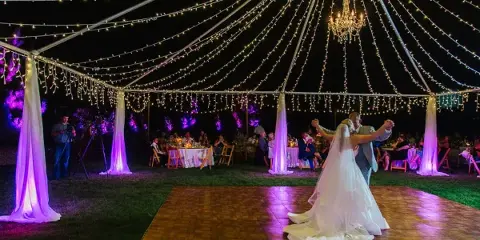 A bride and groom dancing on a wooden dance floor under a canopy of string lights and chandeliers at a wedding reception.