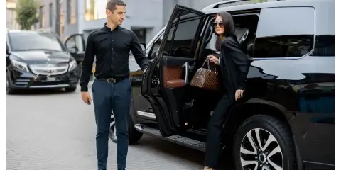 A professional chauffeur in a black shirt and dark trousers holds the door open for a woman exiting a black luxury SUV.
