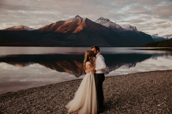 A bride and groom embracing on a rocky shore with a mountain lake in the background.