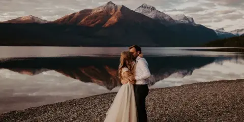 A bride and groom embracing on a rocky shore with a mountain lake in the background.
