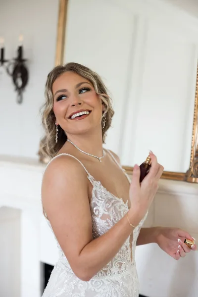 A smiling bride in a white lace wedding dress with styled wavy hair and full makeup, holding a perfume bottle.