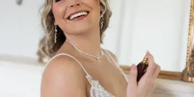 A smiling bride in a white lace wedding dress with styled wavy hair and full makeup, holding a perfume bottle.