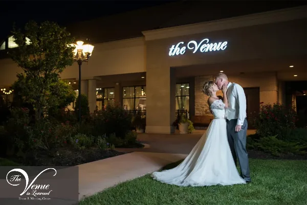 A bride and groom kissing in front of the exterior of The Venue in Leawood at night.