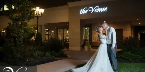 A bride and groom kissing in front of the exterior of The Venue in Leawood at night.