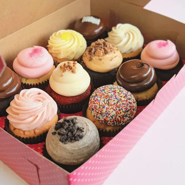 A box filled with an assortment of decorated cupcakes featuring various frostings, sprinkles, and toppings.
