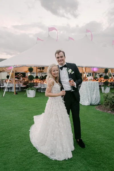 A bride and groom standing on a lawn in front of a large white event tent with pink flags.