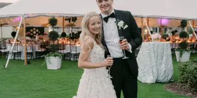 A bride and groom standing on a lawn in front of a large white event tent with pink flags.