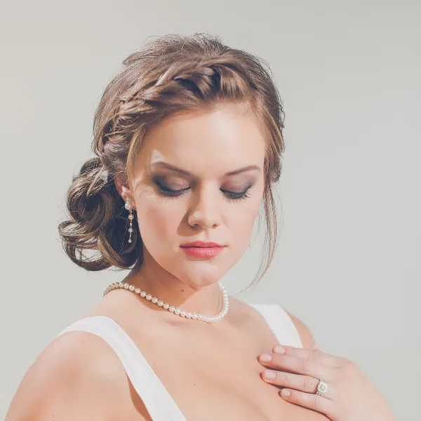 A professional portrait of a bride with an elegant braided updo hairstyle and full makeup, wearing a white dress and pearl accessories.