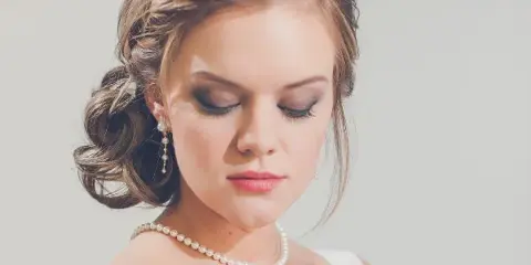 A professional portrait of a bride with an elegant braided updo hairstyle and full makeup, wearing a white dress and pearl accessories.