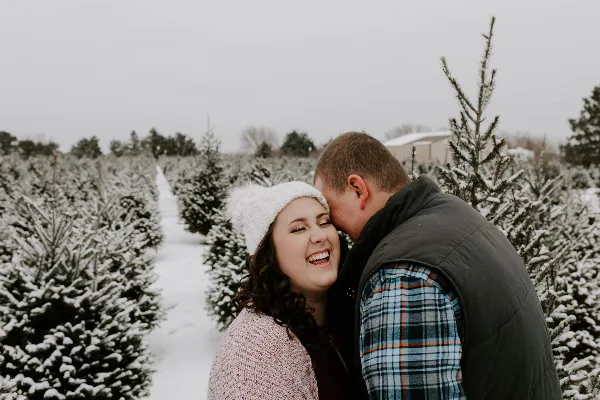 A smiling couple posing together in a snowy Christmas tree farm.