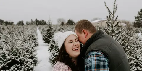 A smiling couple posing together in a snowy Christmas tree farm.