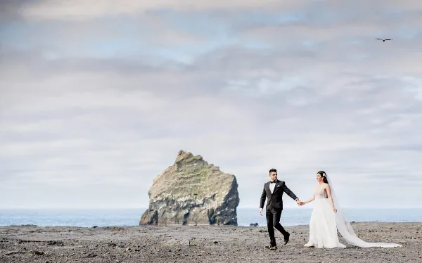 A bride and groom holding hands and walking on a sandy beach with a large rock formation in the background.