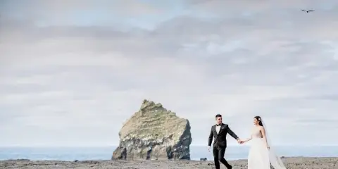 A bride and groom holding hands and walking on a sandy beach with a large rock formation in the background.