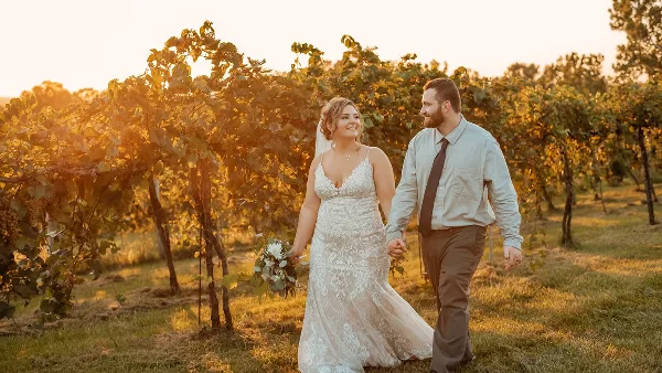 A bride and groom walking hand-in-hand through a vineyard at sunset.