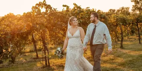 A bride and groom walking hand-in-hand through a vineyard at sunset.